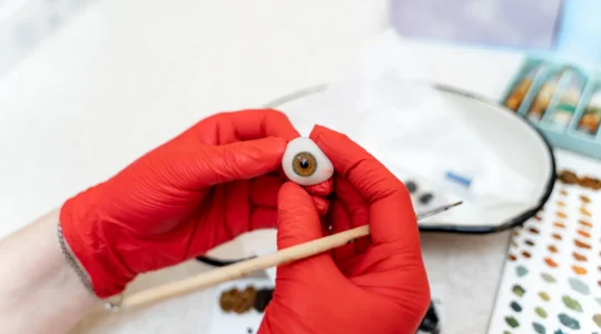 Un oculariste peint à la main l'iris d'une prothèse oculaire sur son établi, sous une lampe loupe.