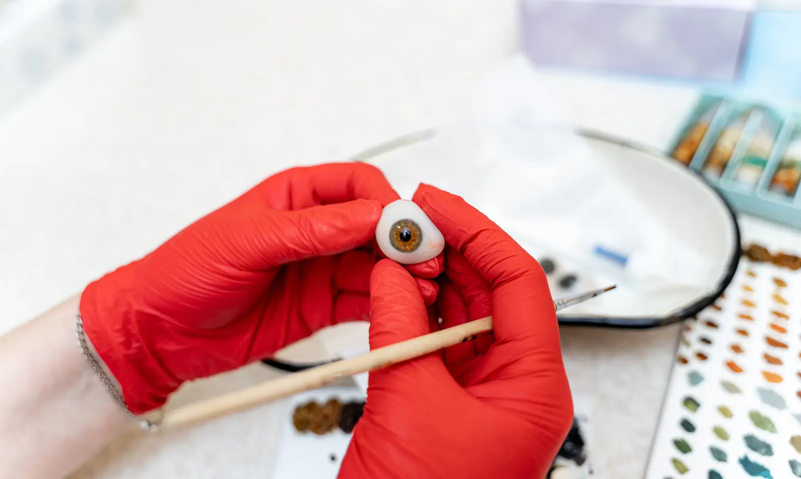 Un oculariste peint à la main l'iris d'une prothèse oculaire sur son établi, sous une lampe loupe.